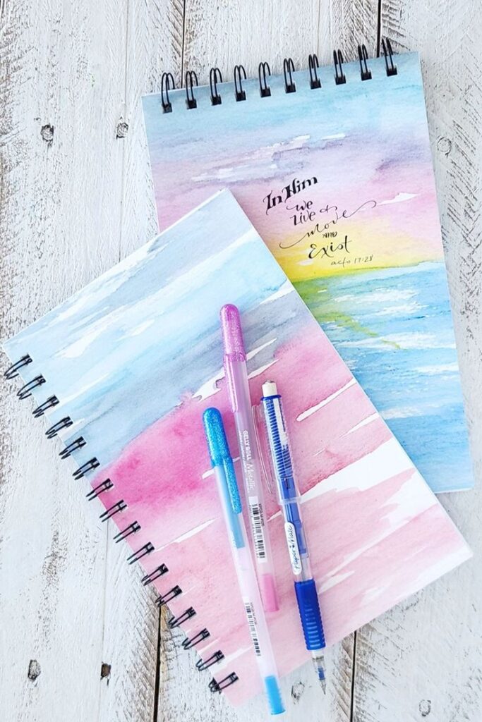 Colorful notebooks with watercolor designs, pens on top, inspirational quote on one cover, wooden background.