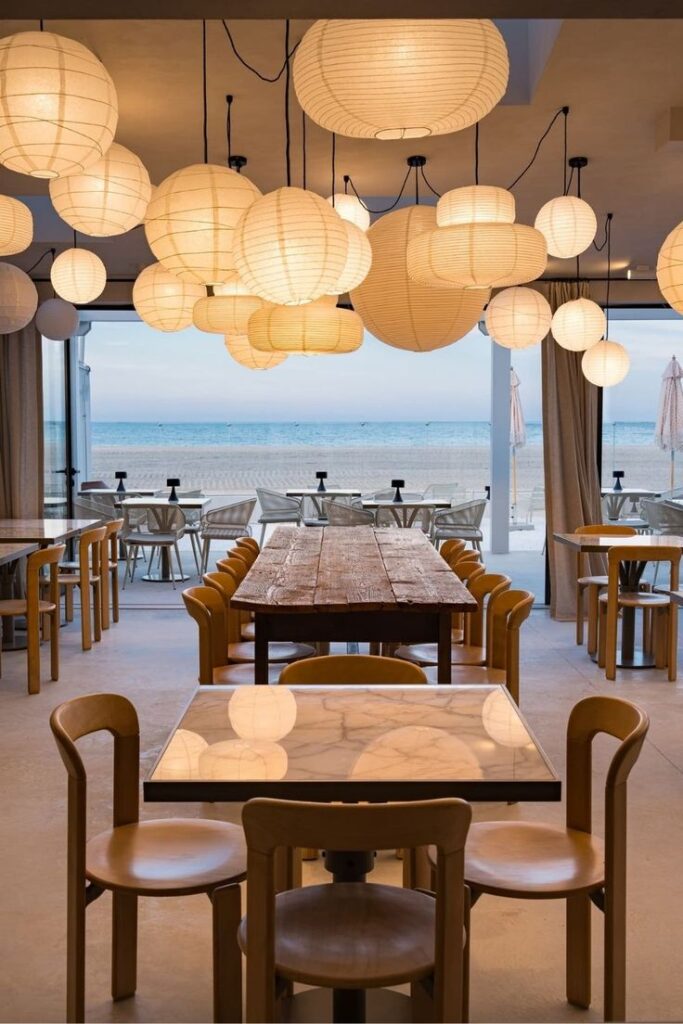 Cozy beachside restaurant interior with wooden tables, chairs, and hanging paper lanterns, overlooking the ocean.