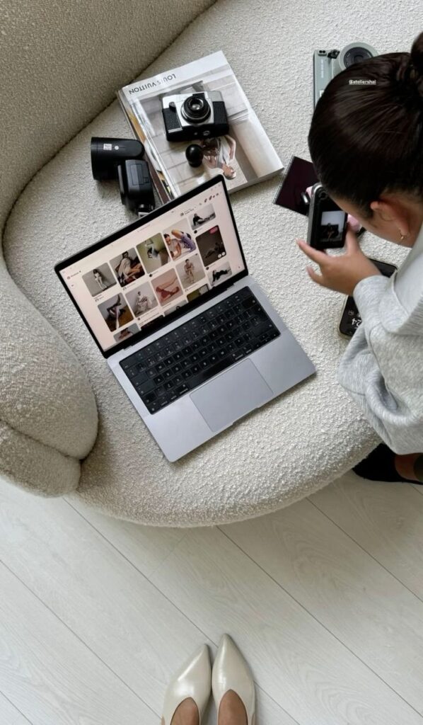 Overhead view of woman using a camera and laptop on cozy beige sofa, surrounded by books and cameras.