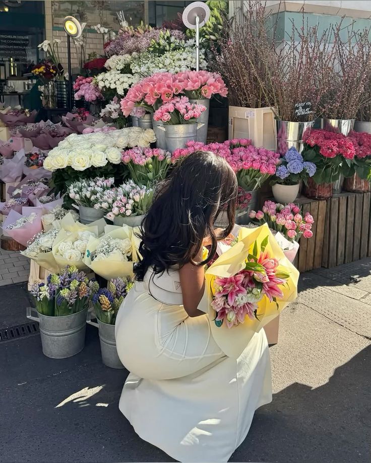 Woman in white dress holding bouquet at a vibrant outdoor flower market, surrounded by colorful blooms.