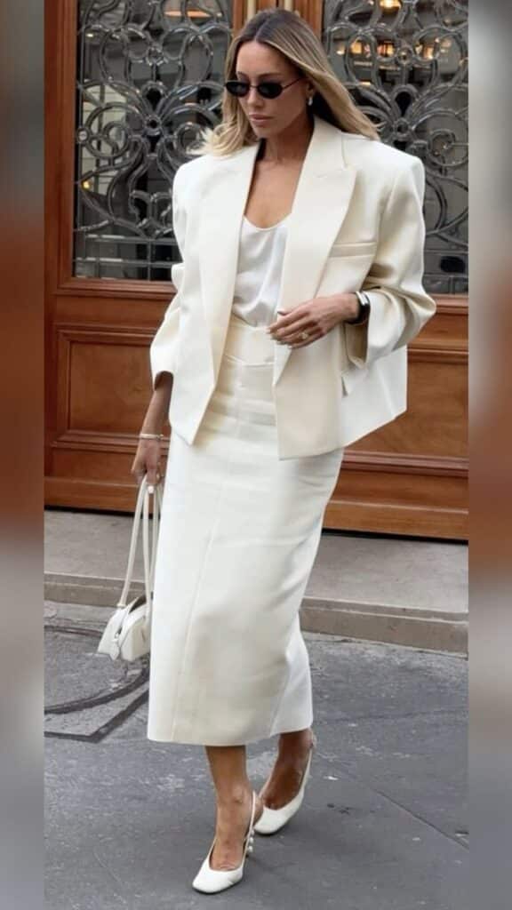 Stylish woman in white blazer and skirt, sunglasses, and heels, walking confidently with a matching handbag.