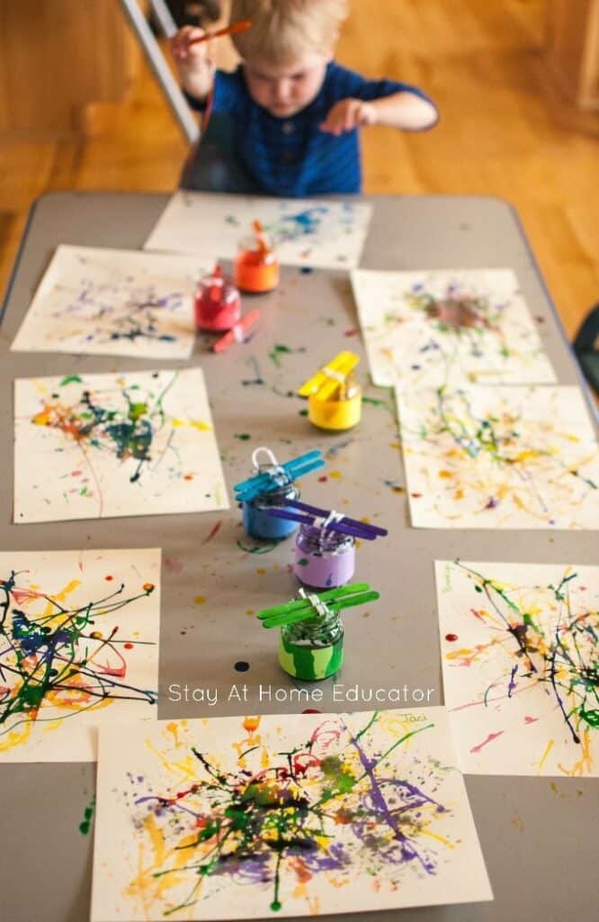 Child painting with colorful splatter art on paper at a table with paint jars.