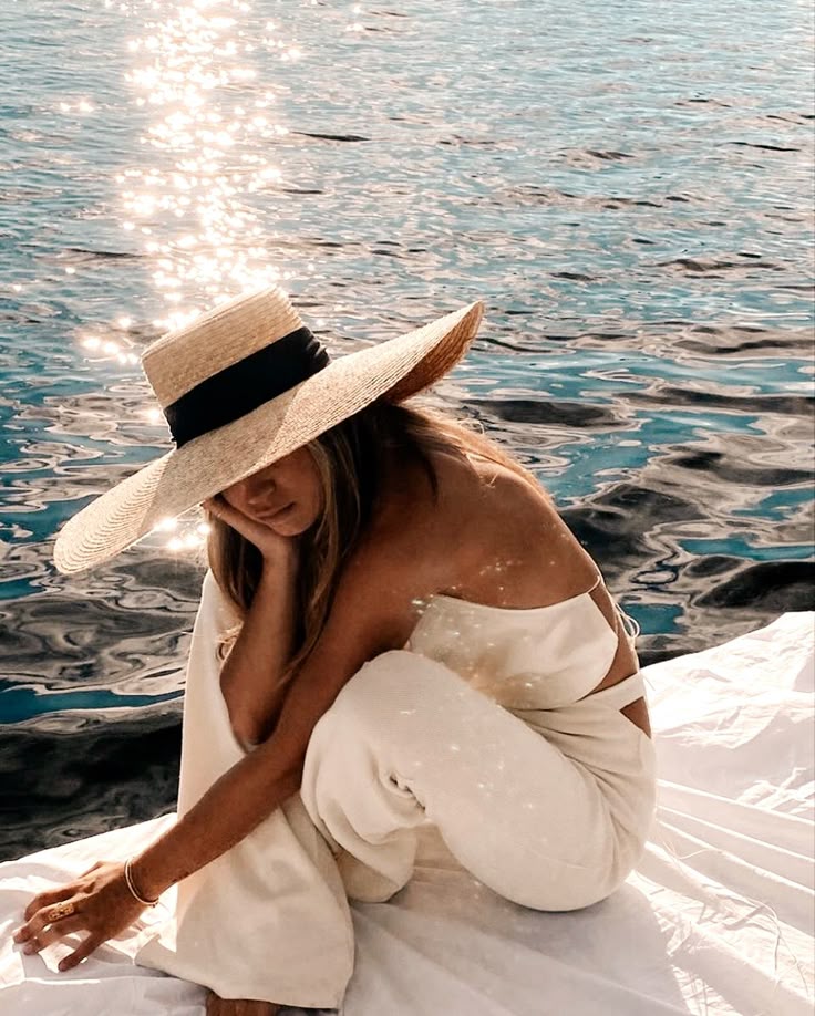 Woman in wide-brim hat and white outfit sitting by sparkling water, enjoying a serene day outdoors.
