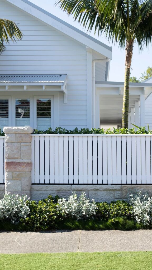 Modern coastal home with white siding, palm tree, and neatly landscaped front yard and fence in bright sunlight.