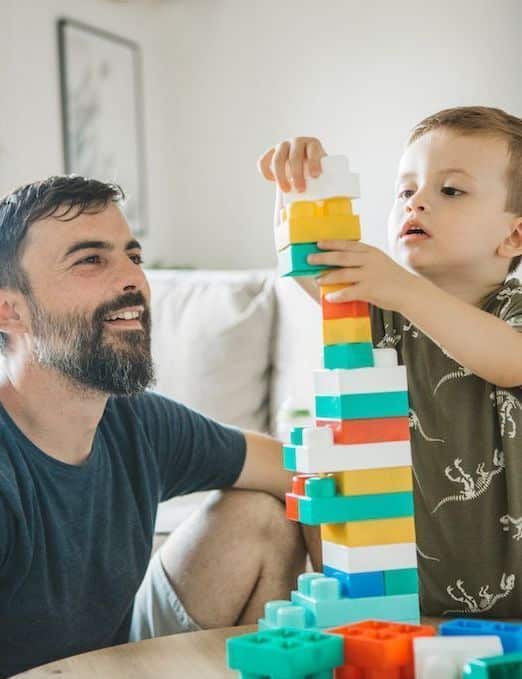 Father and child building a colorful tower with blocks at home, smiling and bonding.