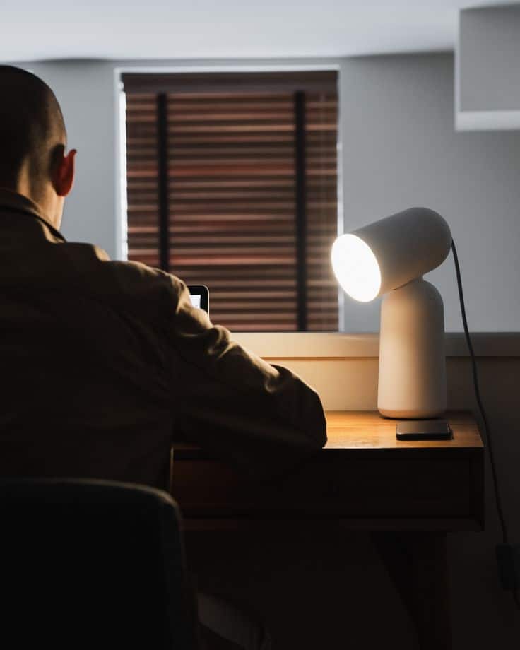 Person working at a desk with a modern lamp, focused on a laptop screen in a dimly lit room.