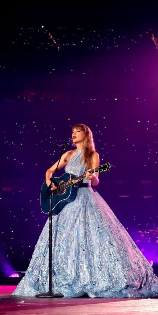 Singer in a sparkling blue dress performs on stage with a guitar under purple lights.