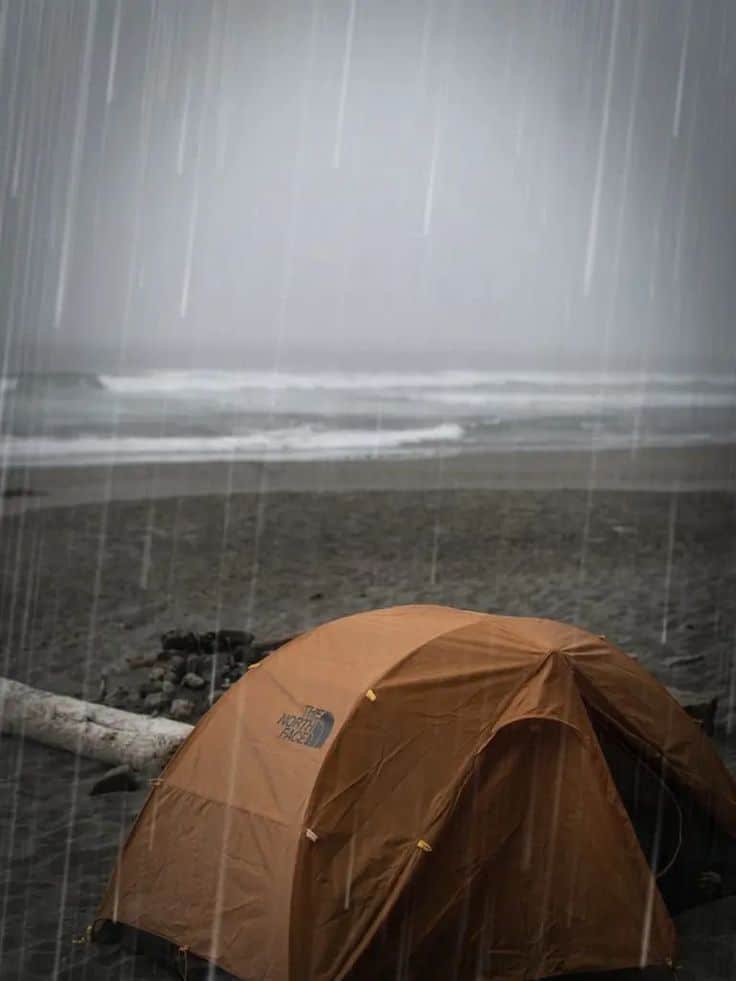 Orange tent on a rainy beach with ocean waves in the background, conveying a moody outdoor adventure scene.