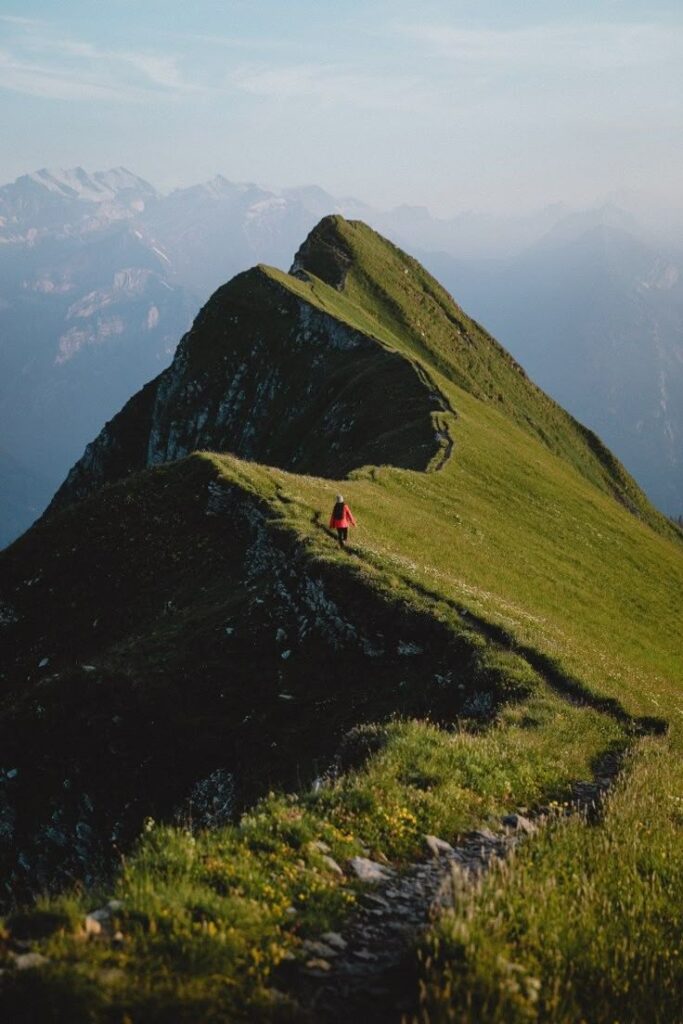 A person in a red jacket hikes along a scenic mountain ridge with a beautiful landscape view in the background.