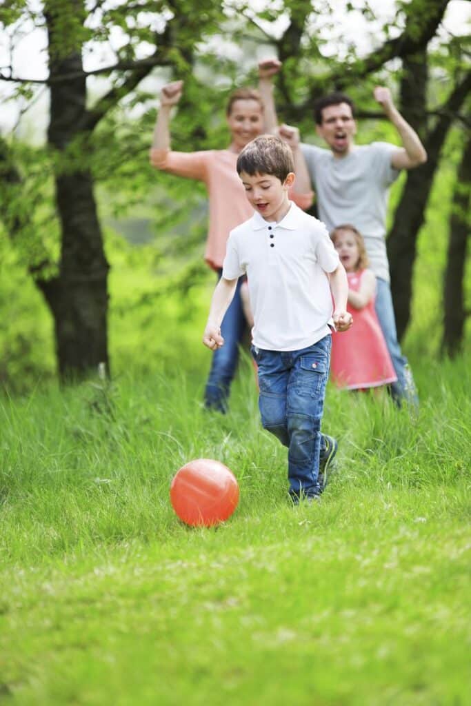 Child playing with orange ball in park, family cheering in the background amidst green trees and grass.