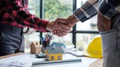 Two people shaking hands over construction plans and model house, symbolizing real estate or building agreement.