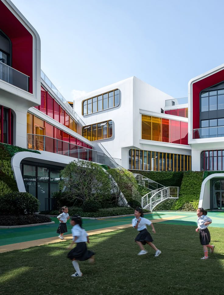 Modern school building with colorful windows, children playing outside on green lawn under clear blue sky.