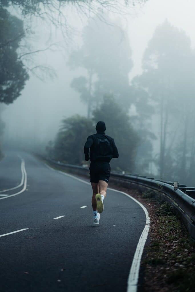 Jogger running on a foggy road surrounded by tall trees, creating a serene and moody atmosphere.
