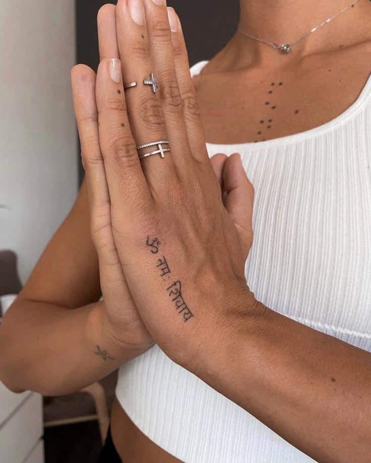 Woman in white top with Namaste hands, showcasing a Sanskrit tattoo and rings on fingers.