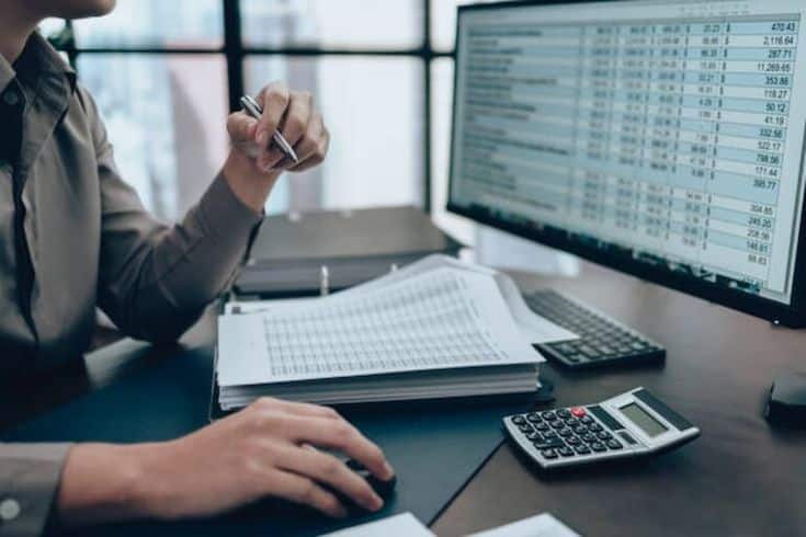 Person analyzing financial data on a computer screen, with spreadsheets and a calculator on the desk.