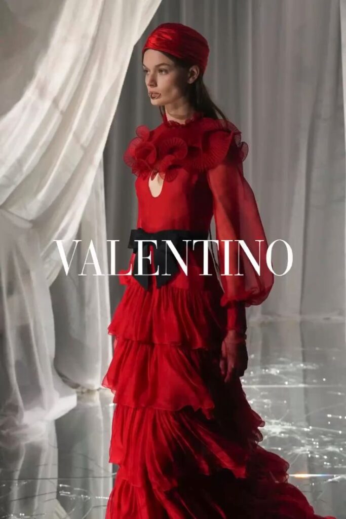 Model in elegant red Valentino gown with ruffled details, posing gracefully indoors against sheer curtains. Fashion Design