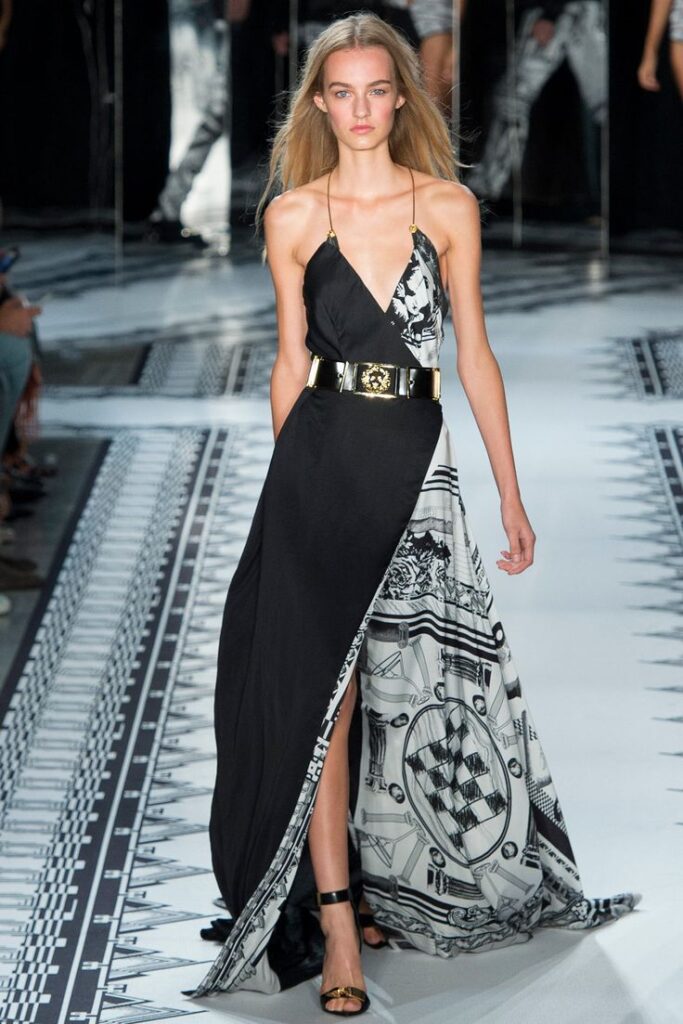 Model in black and white geometric print gown on runway, showcasing elegant fashion design with gold belt accent.