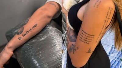 Close-up of two arms with various tattoos, featuring text and small designs, on a blurred background.