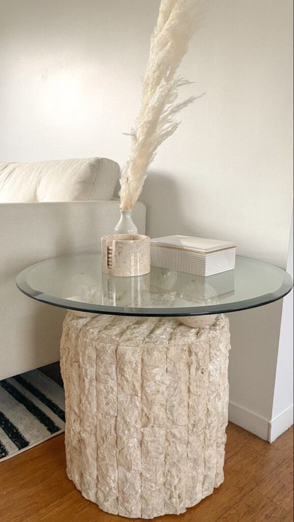 Modern side table with glass top, pampas grass vase, and textured base in a cozy living room setting.
