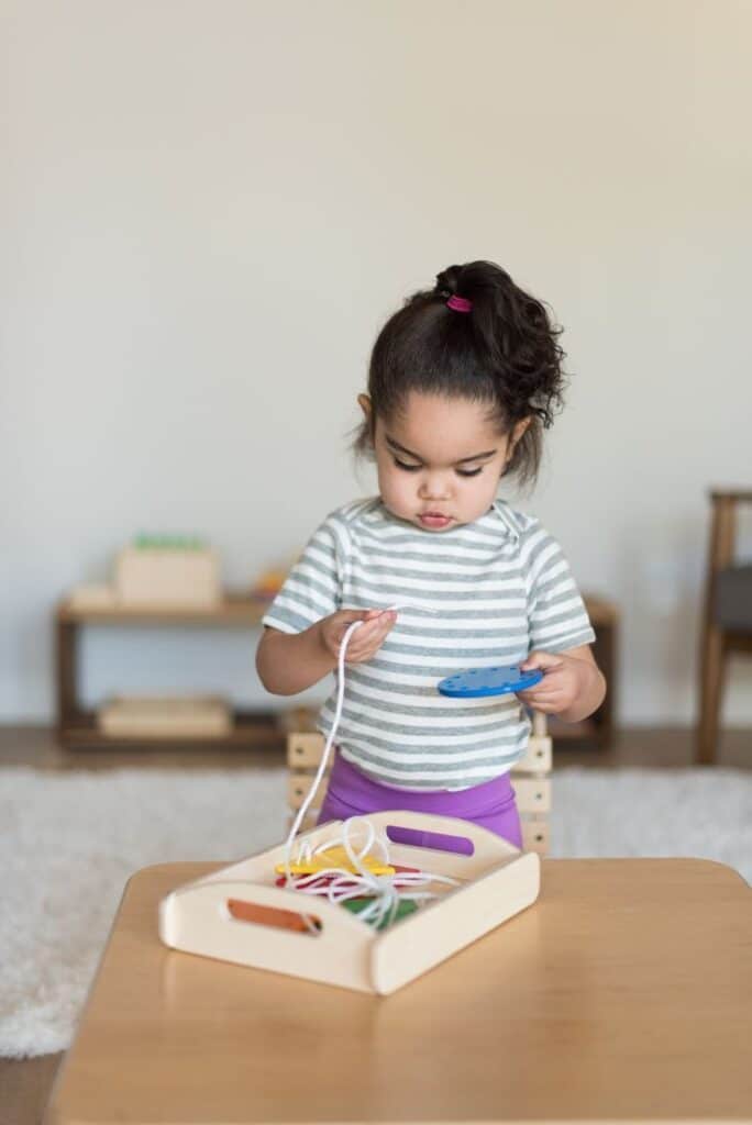 Vor der Vorschule gibt es Monti Kids_ Toddler engaged in threading activity with colorful strings and board at home. | Sky Rye Design Toddler engaged in threading activity with colorful strings and board at home.