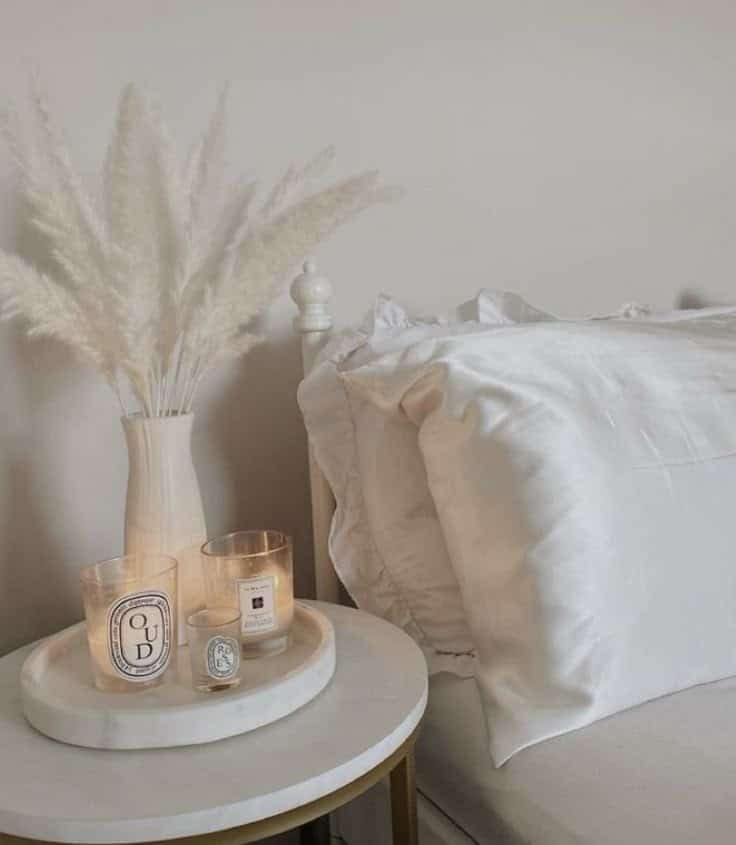 Elegant bedroom with white bed linens, pampas grass in vase, and scented candles on a stylish nightstand.