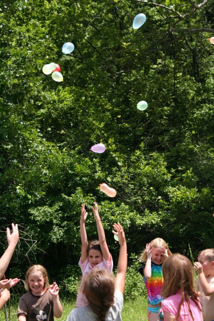 Children enjoying a water balloon fight outdoors on a sunny day, surrounded by green trees.