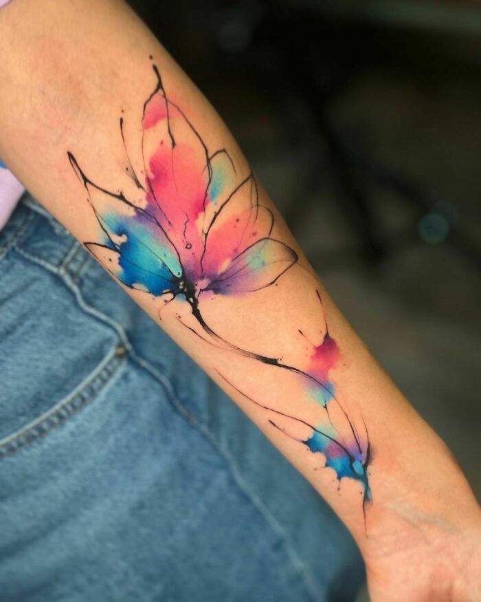 Vibrant watercolor butterfly tattoo on forearm, featuring splashes of pink, blue, and purple ink for a dynamic look.