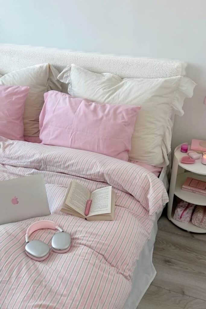 Cozy bedroom with pink bedding, open book, headphones, and laptop on bed, creating a relaxing atmosphere.