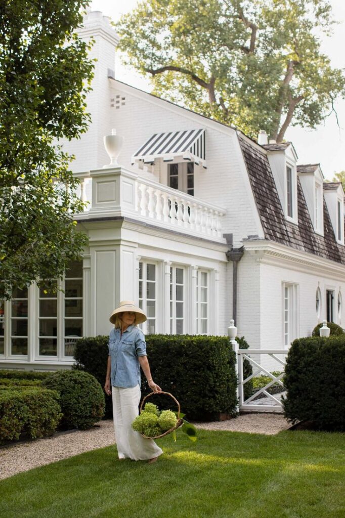 Woman in garden with straw hat and basket of hydrangeas, near elegant white house with green landscaping.