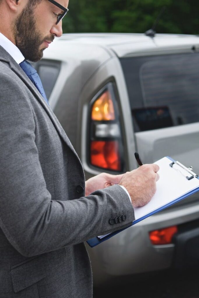 What Happens When Car Insurance Denies a Claim_ Man in suit inspecting vehicle while writing on clipboard outdoors. | Sky Rye Design Man in suit inspecting vehicle while writing on clipboard outdoors.