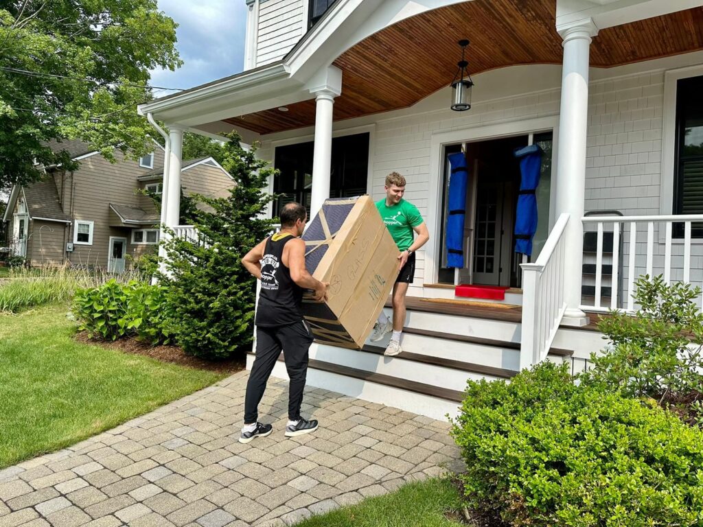 Two people carrying a large box into a house with a porch, symbolizing teamwork and moving.