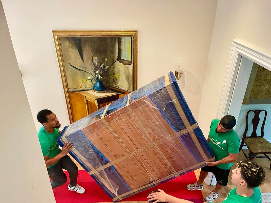 Movers carefully handling a large, wrapped item in a hallway with artwork on the wall.