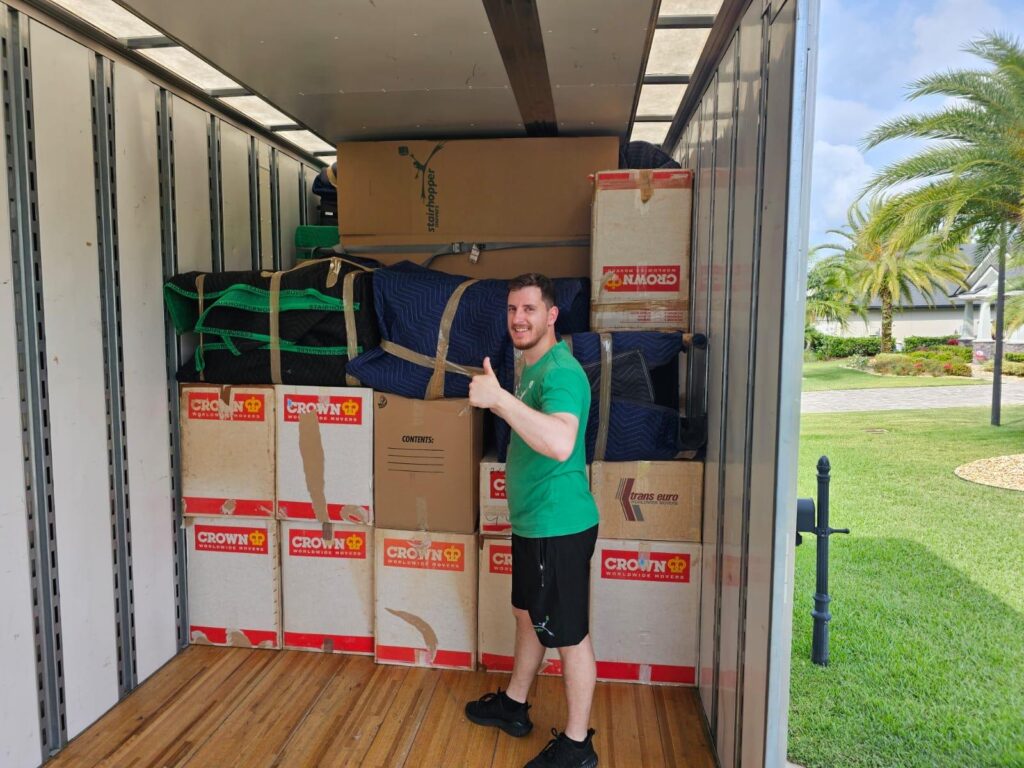Man gives thumbs up in moving truck filled with packed boxes and furniture blankets, ready for relocation.