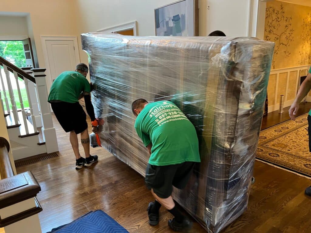 Professional movers carefully transporting a wrapped couch in a home with hardwood floors.