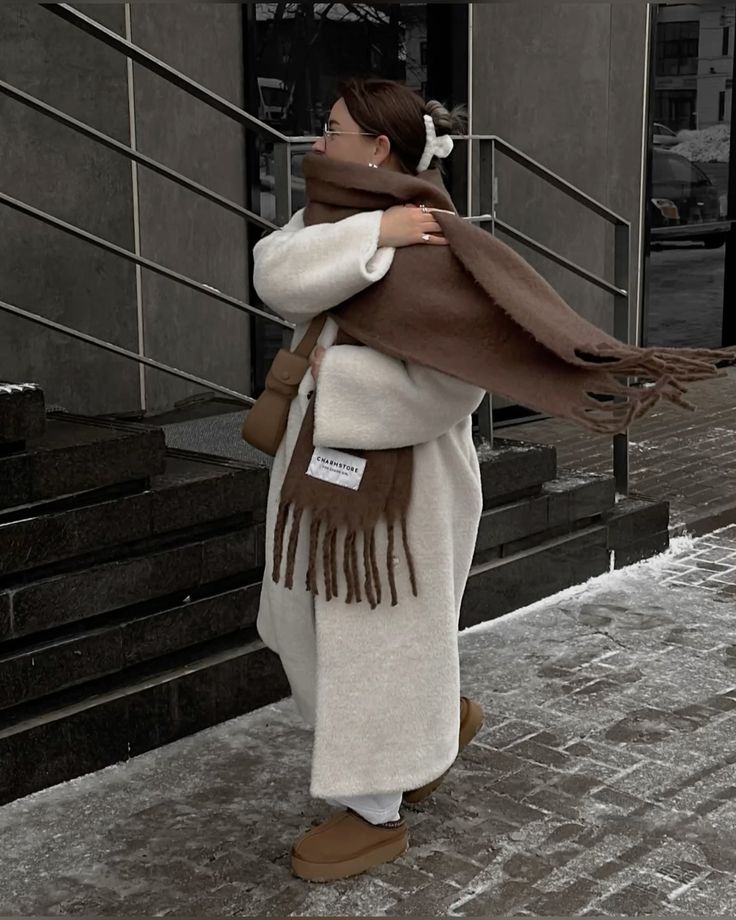 Person in cozy winter outfit with oversized scarf and coat walking on snowy city sidewalk.