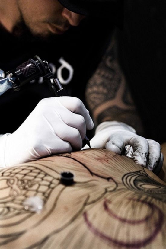 Tattoo artist inking a detailed design on skin, wearing gloves and using a tattoo machine in a studio setting.