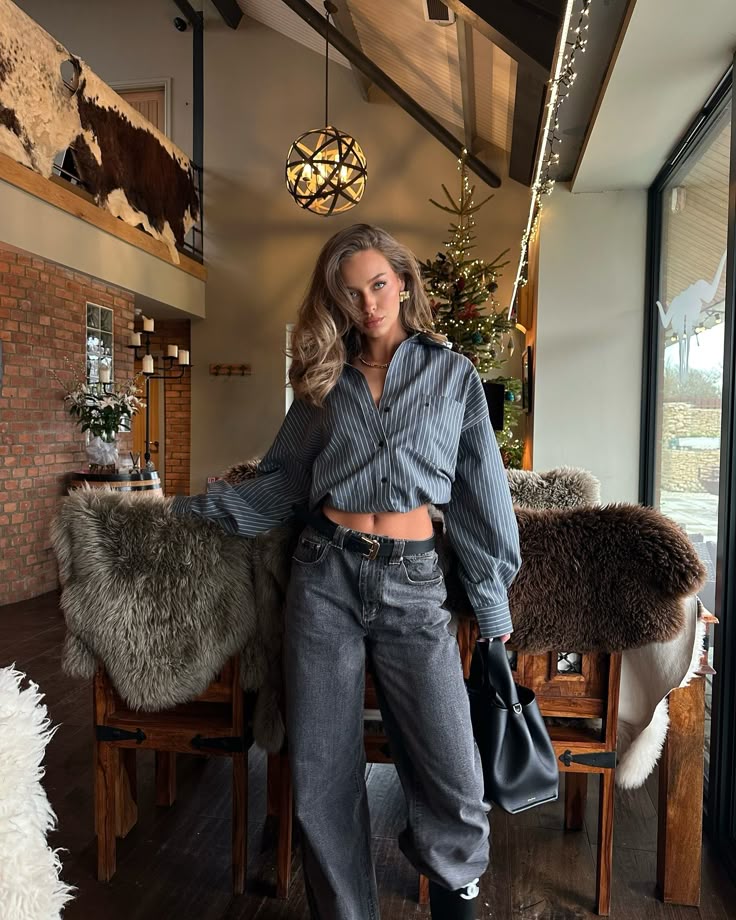 Stylish woman in casual attire poses indoors with Christmas tree and rustic decor.