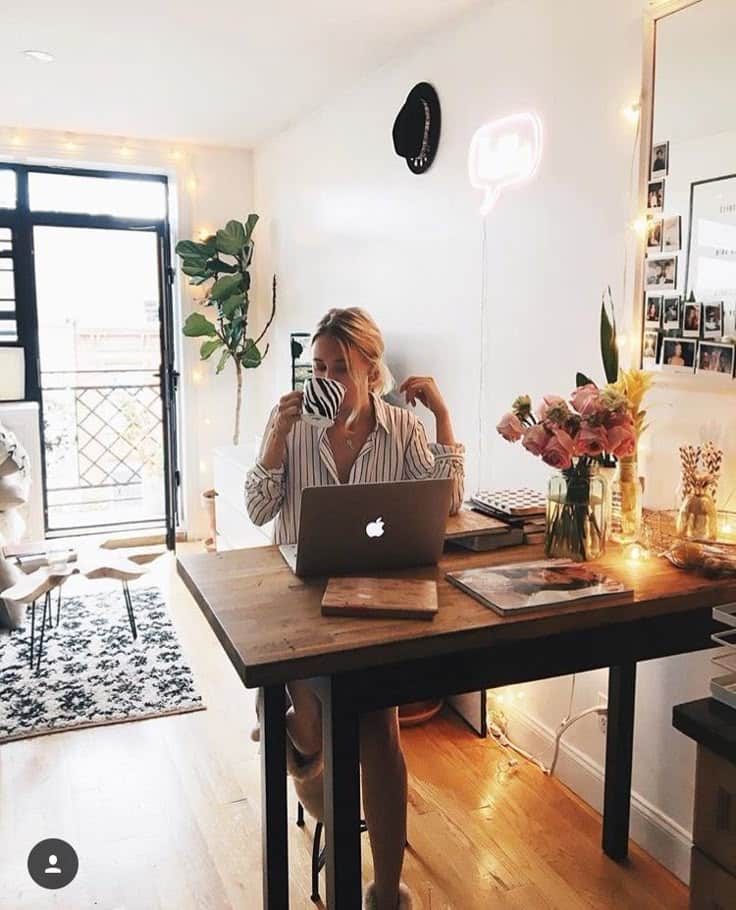 Cozy home office with person sipping coffee at desk, laptop open, surrounded by plants and decor, soft lighting.
