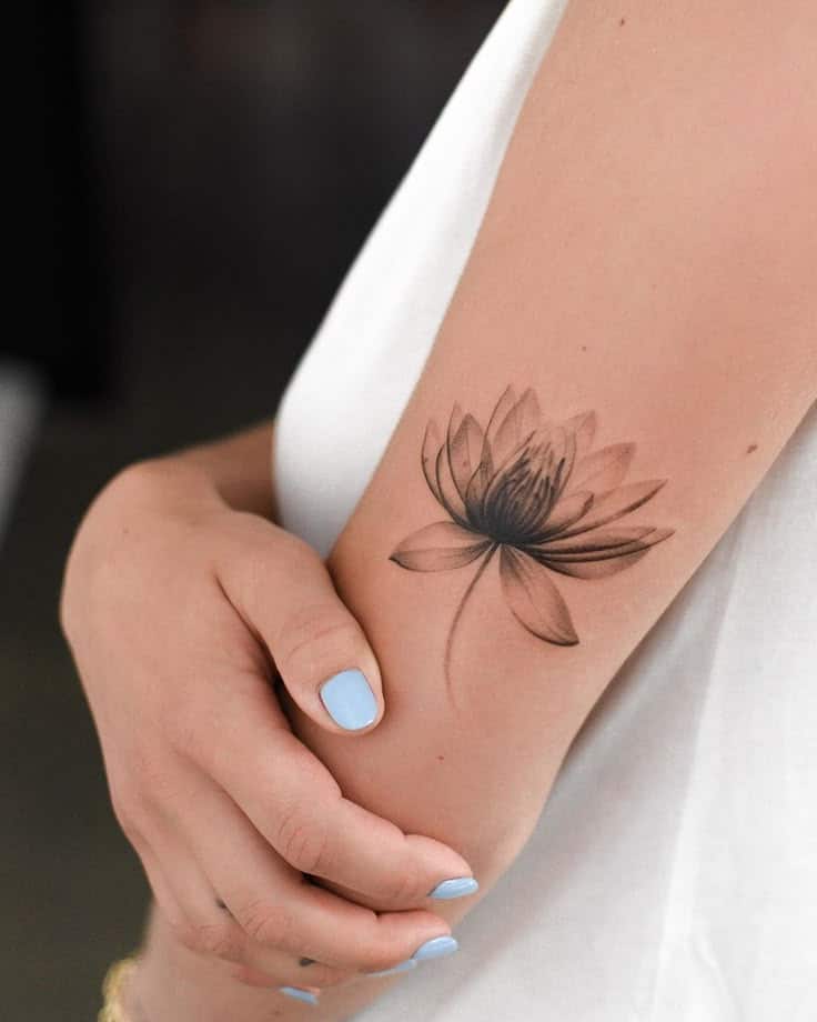 Delicate lotus flower tattoo on forearm, held by person with light blue nails and a white top.