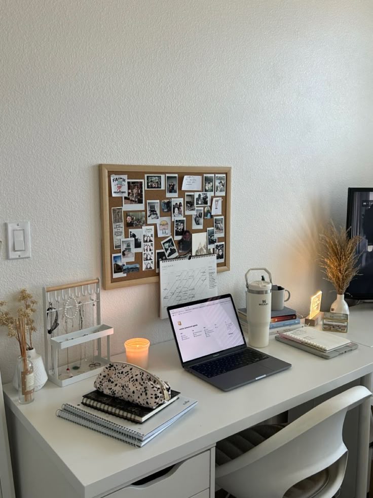 Cozy home office with laptop, candle, and decor on a white desk. Bulletin board displays photos and notes on the wall.