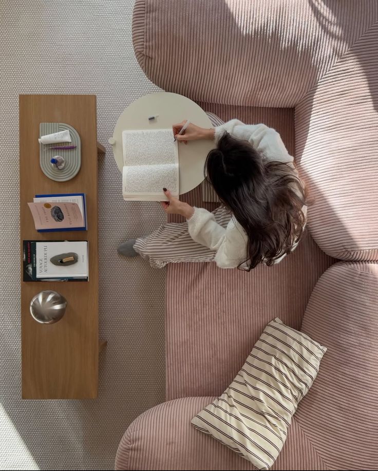 Woman writing in a book on a cozy pink sofa, overhead view, with books and decor on a wooden table nearby.