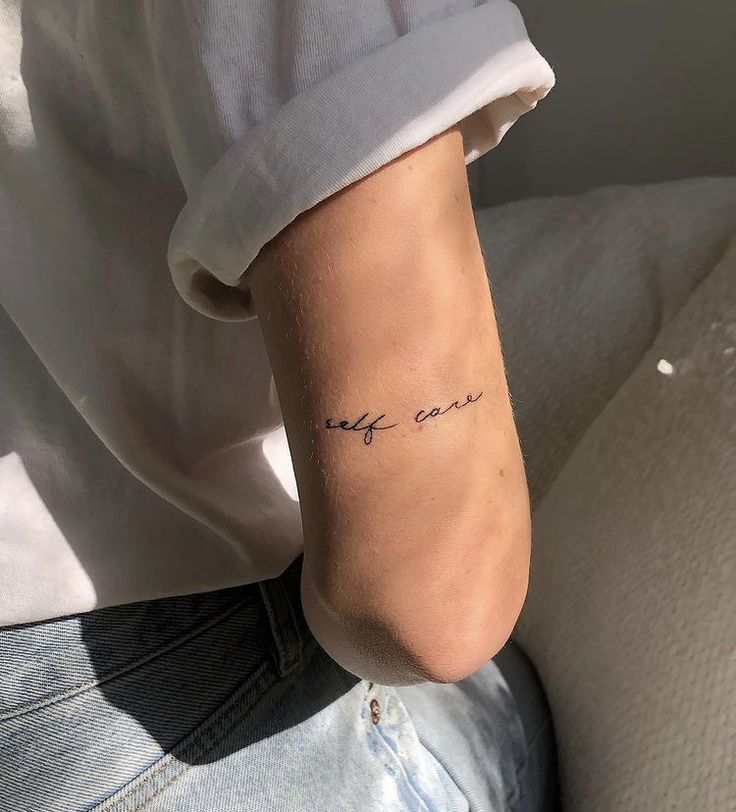 Close-up of arm with self care tattoo in delicate script, wearing white shirt and jeans, sunlight on skin.