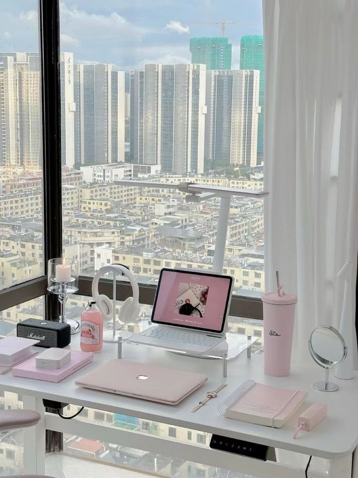 Chic home office setup with pink accessories and laptop, overlooking city skyline through large window.