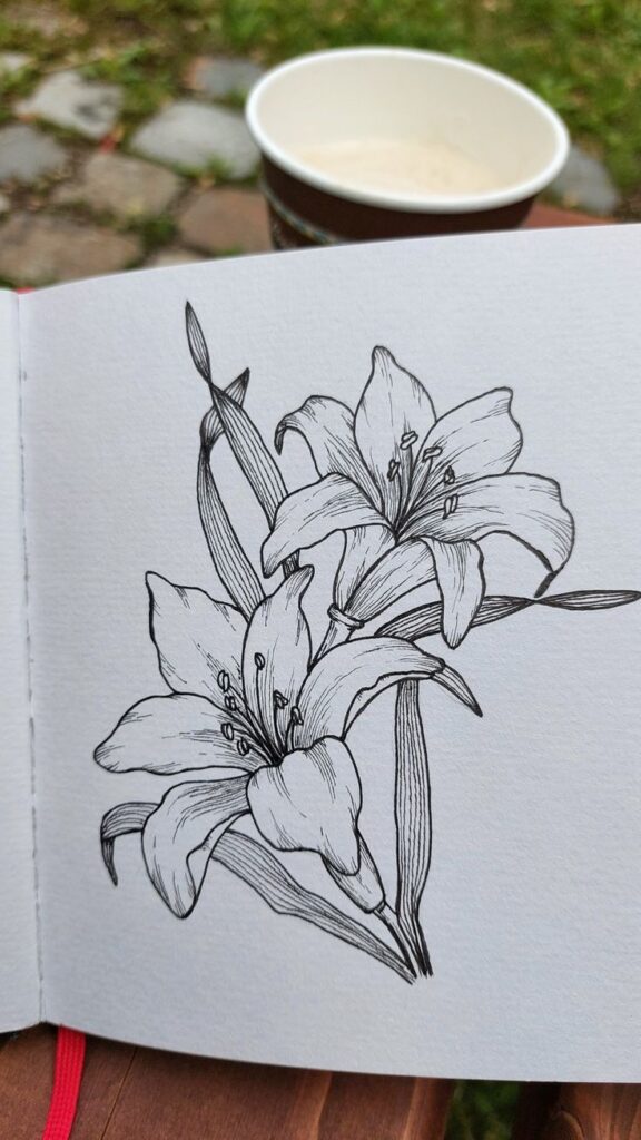 Black and white lily sketch in a notebook with a cup of coffee on a wooden table outdoors.