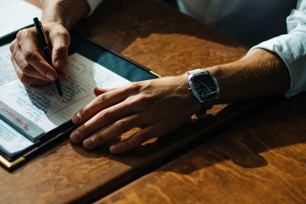 adolfo-felix-Yi9-QIObQ1o-unsplash Person writing in a notebook at a wooden desk, wearing a wristwatch, in natural light. | Sky Rye Design Person writing in a notebook at a wooden desk, wearing a wristwatch, in natural light.