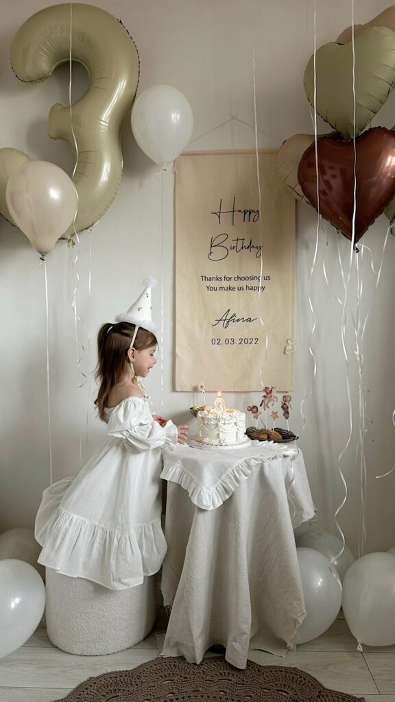 Girl in white dress at birthday party with cake and balloons, celebrating her third birthday.