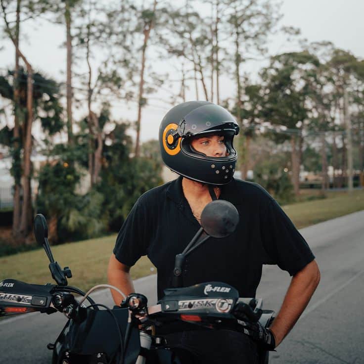 Man in black moto helmet on motorcycle, roadside with trees in background.