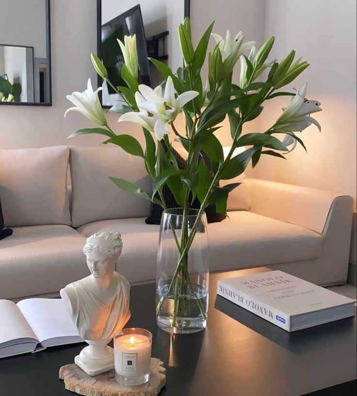 Elegant living room with white lilies in a vase, candle, book, and sofa for a cozy ambiance.