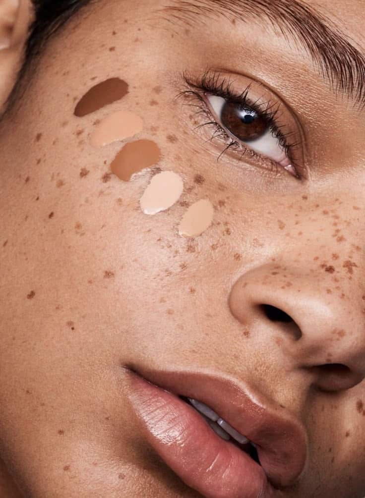 Close-up of a person's face with varied makeup foundation swatches and natural freckles.
