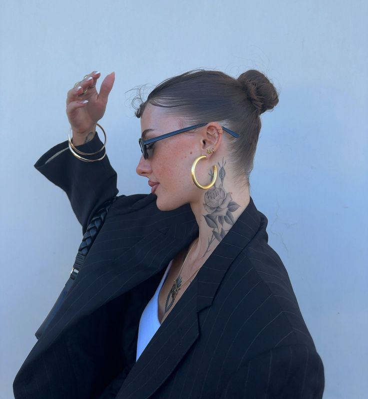 Trendy woman with a floral neck tattoo, large hoop earrings, and sunglasses in a stylish black blazer.
